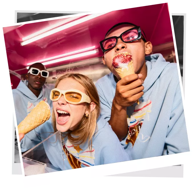 Two people in sunglasses eating messy ice cream cones with a third person in the background.